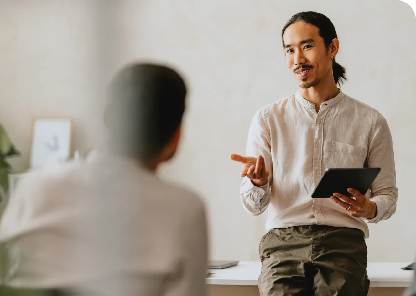 Man with a tablet gestures while sitting on a desk, engaging in conversation with another person in a blurred foreground.