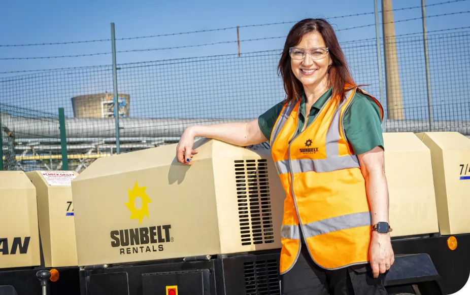 Woman in an orange safety vest and glasses smiles, standing next to Sunbelt Rentals equipment, with industrial background.