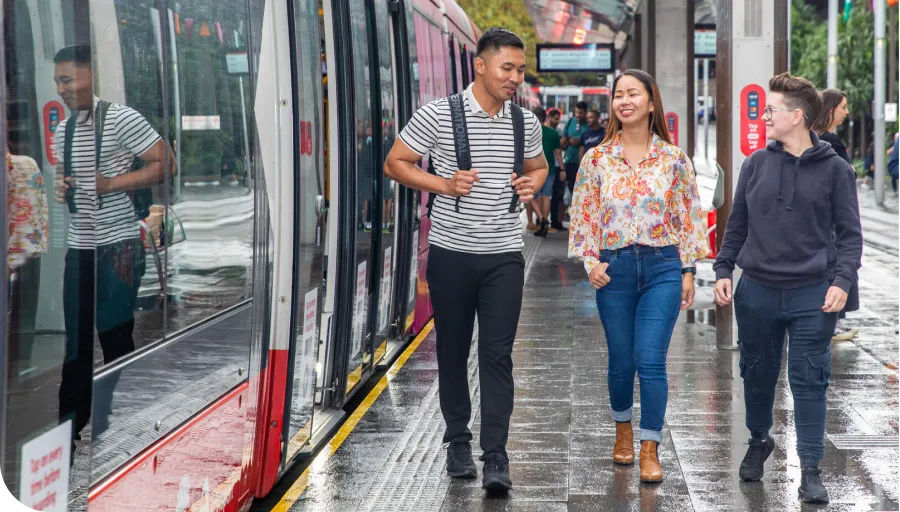 Three people walking on a wet tram platform, smiling and chatting. A tram is parked beside them.