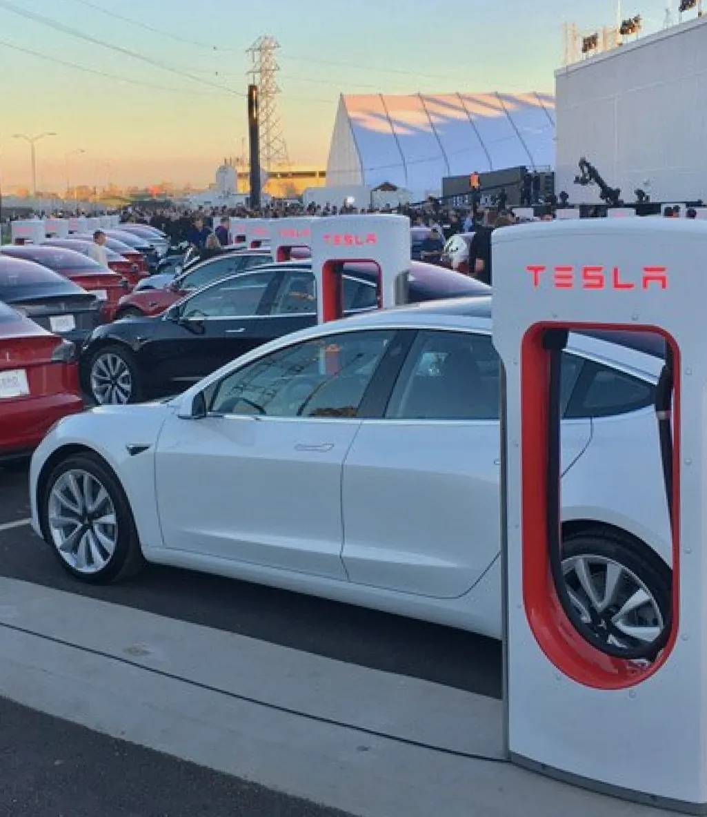 A white Tesla car is parked and charging at a Tesla Supercharger station, with several other Tesla vehicles in the background.