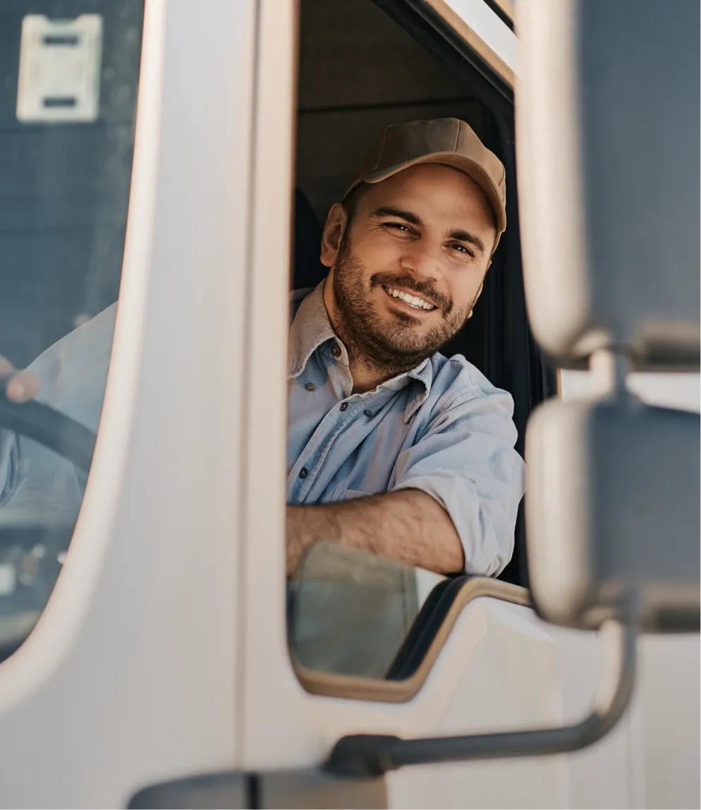 Smiling man in a cap and shirt sitting in a truck driver's seat, looking out the open window, with a side mirror visible.