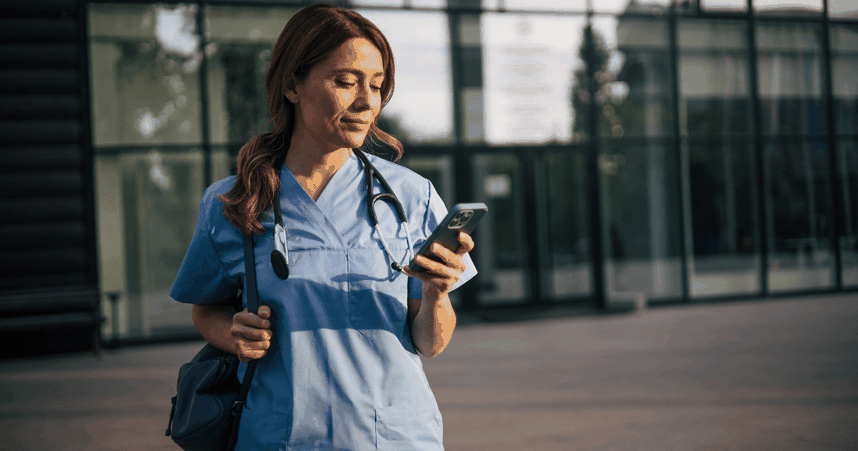 A healthcare professional in blue scrubs and a stethoscope checks her phone while standing outside a modern building.