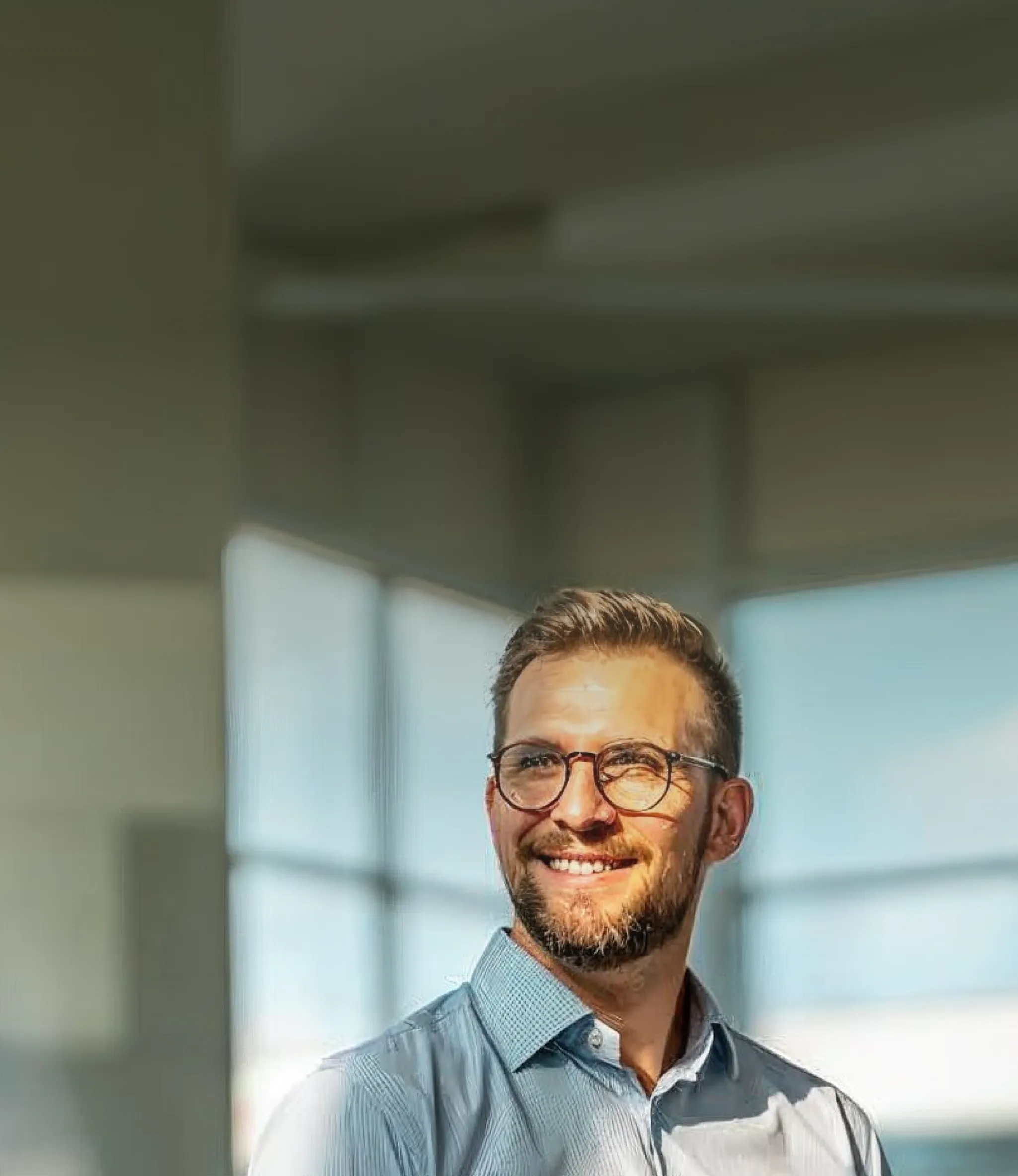 A man with glasses and a beard smiles while standing indoors, wearing a light blue shirt. Bright windows are in the background.