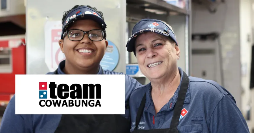 Two smiling employees in Domino's uniforms stand together in a kitchen, with a "Team Cowabunga" logo in the foreground.