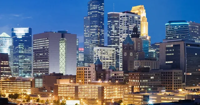 A vibrant city skyline at dusk with illuminated skyscrapers and a historic clock tower, set against a deep blue sky.