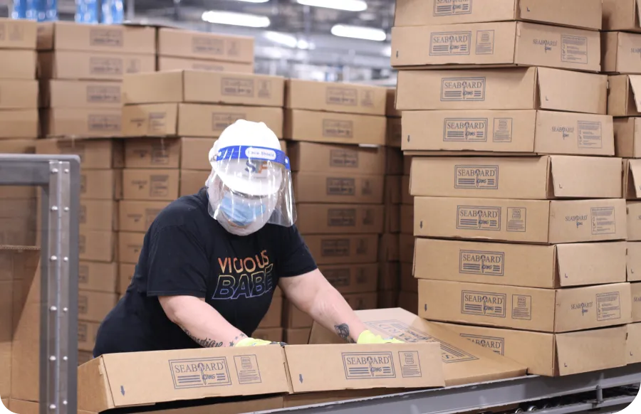 Worker in protective gear stacks cardboard boxes labeled "Seaboard" in a warehouse, surrounded by tall stacks of similar boxes.
