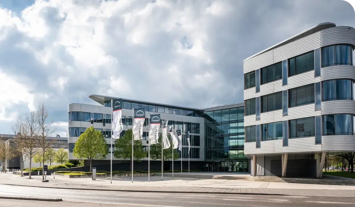 Modern office building with glass facade, surrounded by trees and flags, under a partly cloudy sky.