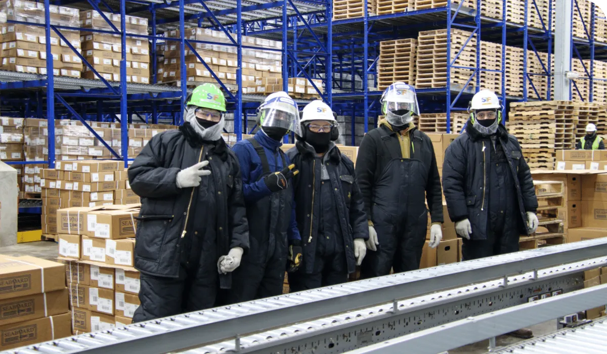 Five workers in protective gear and helmets stand in a warehouse with stacked boxes and shelves, posing for a group photo near a conveyor belt.