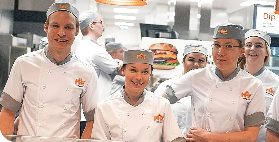 Group of smiling fast-food employees in uniform behind a counter, with a burger poster in the background.