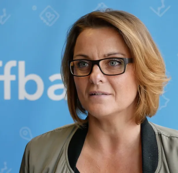 A woman with glasses and short hair stands in front of a blue backdrop with white text.