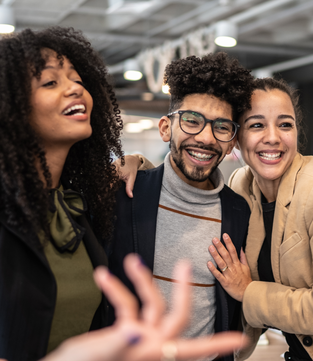 Three people smiling and embracing in a modern indoor setting, with one person gesturing playfully towards the camera.