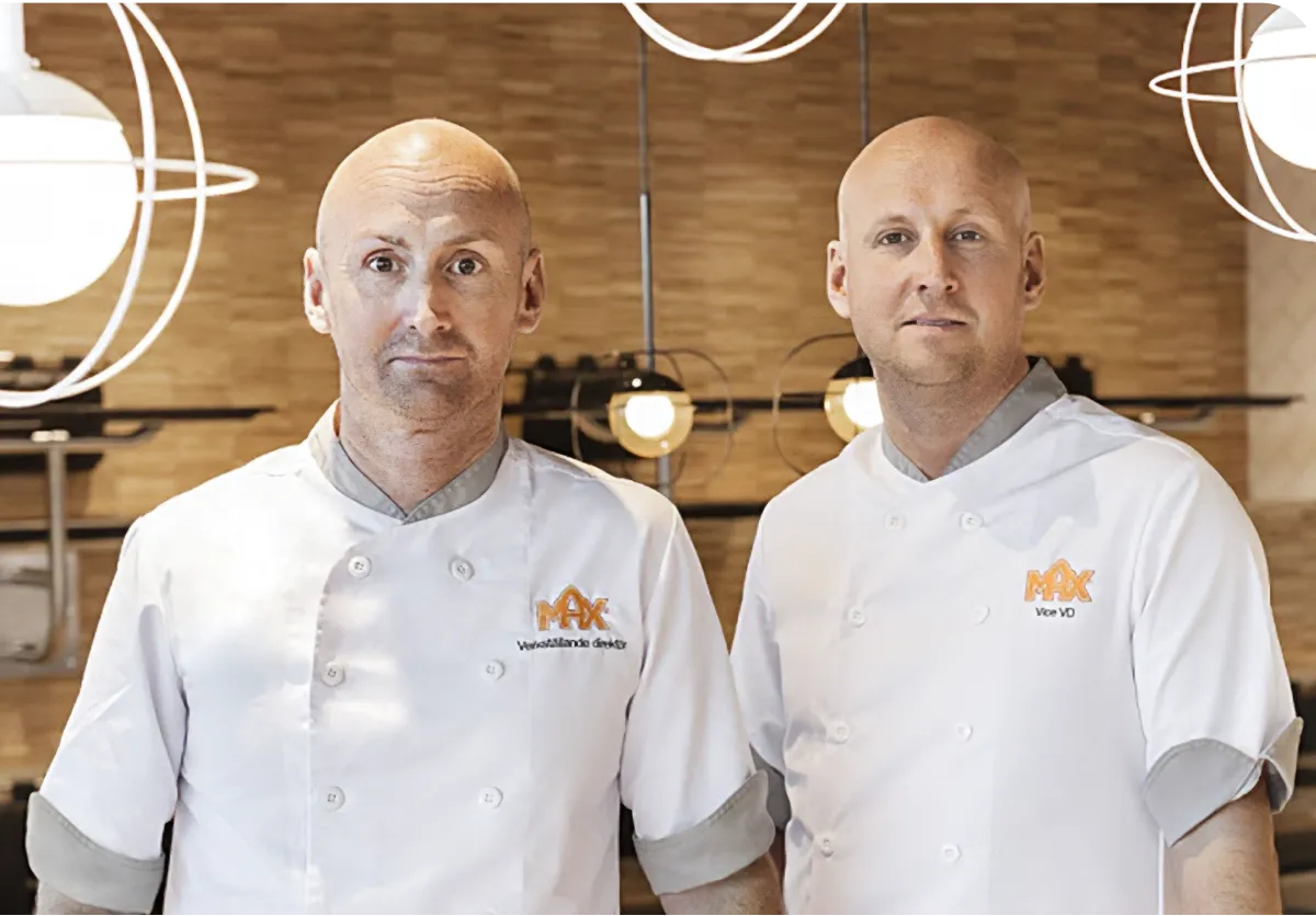Two chefs in white uniforms stand in a modern kitchen with wooden accents and round pendant lights.