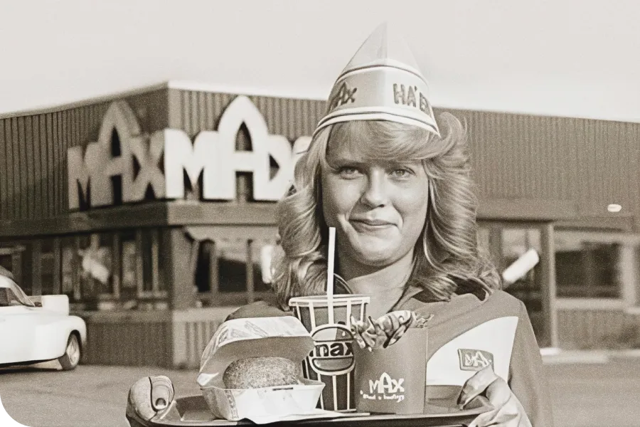Smiling woman in a vintage fast-food uniform holds a tray with a burger, fries, and drink in front of a Max restaurant.