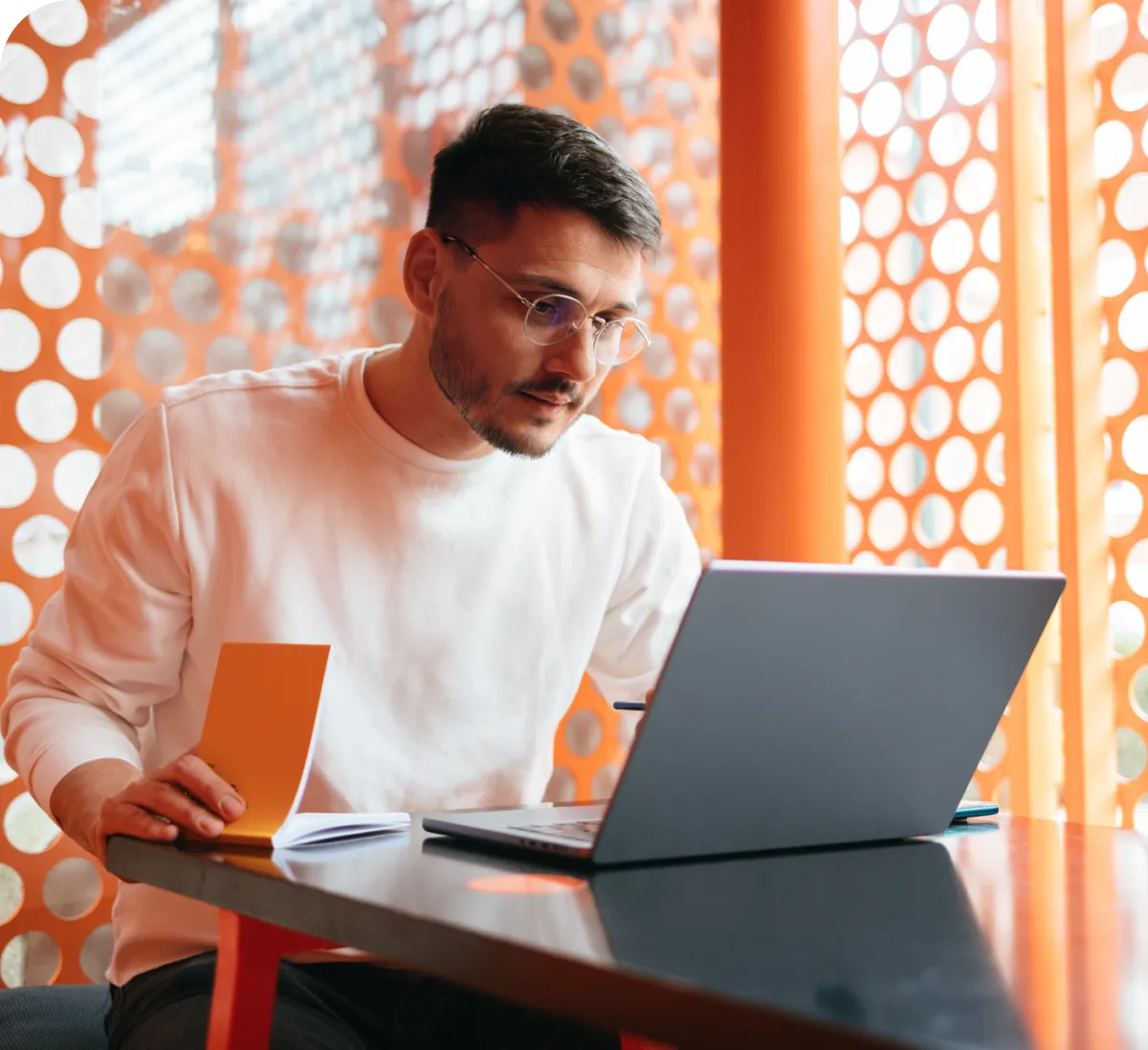 Focused man working on a laptop in a modern, orange-themed workspace, holding a yellow notebook.