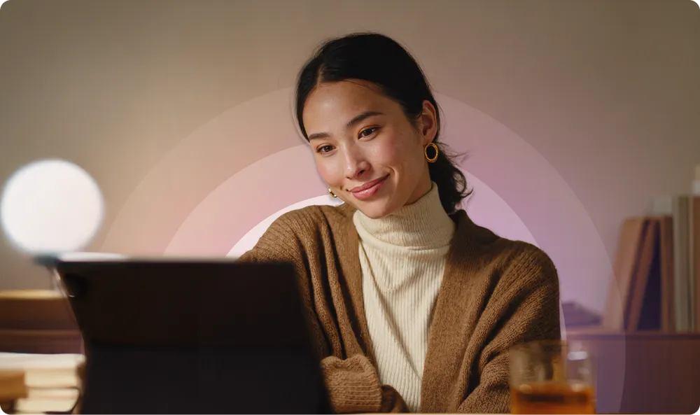 Woman in a cozy sweater smiles while looking at a tablet, with a warm light and books in the background.