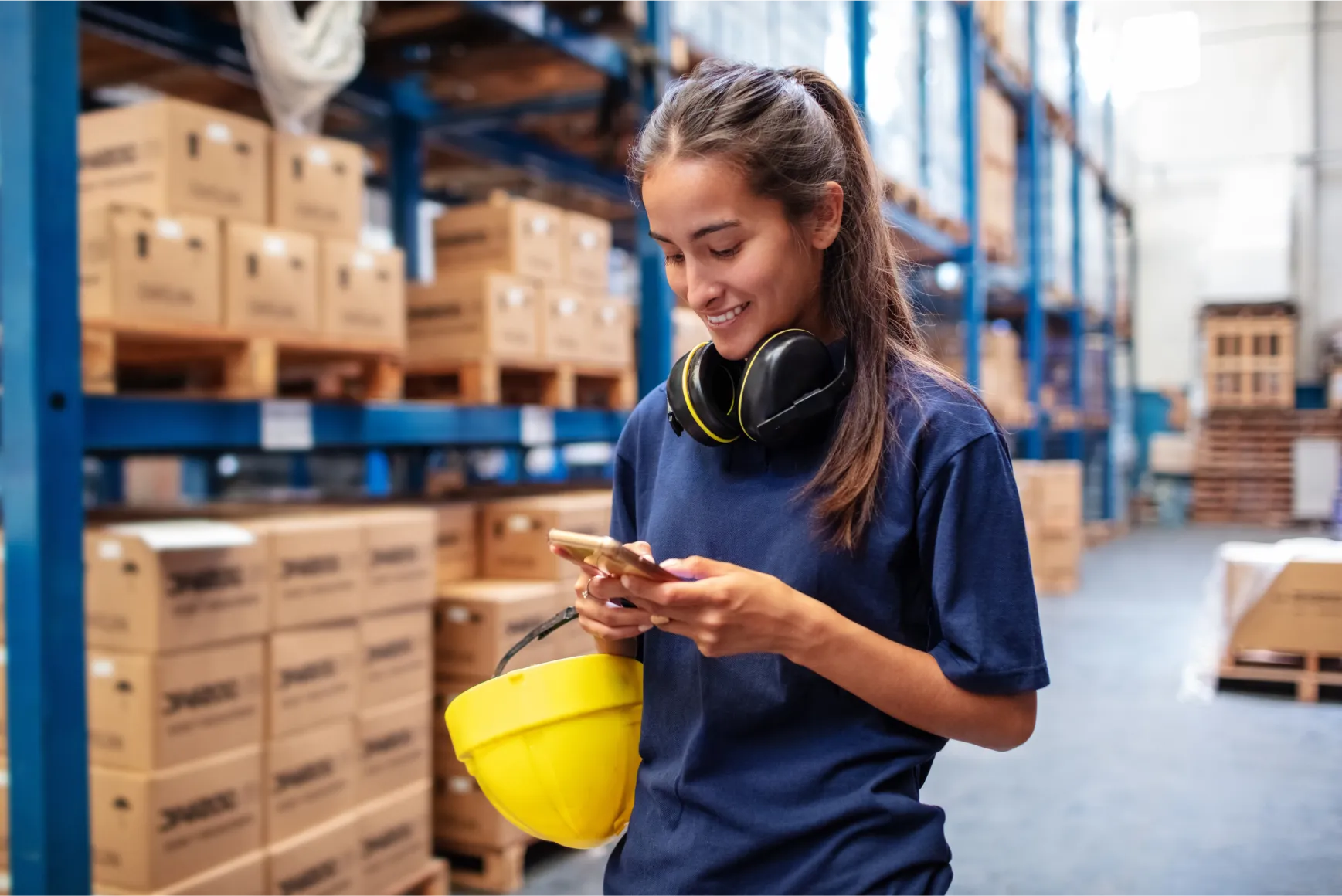 Woman in a warehouse wearing a blue shirt, holding a yellow hard hat, and using a smartphone. Shelves with boxes are in the background.