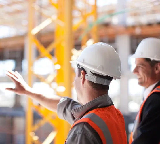 Two construction workers in safety gear and helmets discuss a project on-site, with scaffolding and equipment in the background.