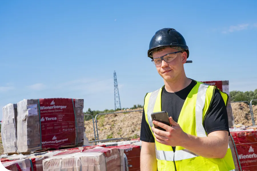 Construction worker in a hard hat and safety vest using a smartphone at a building site with brick stacks and blue sky in the background.
