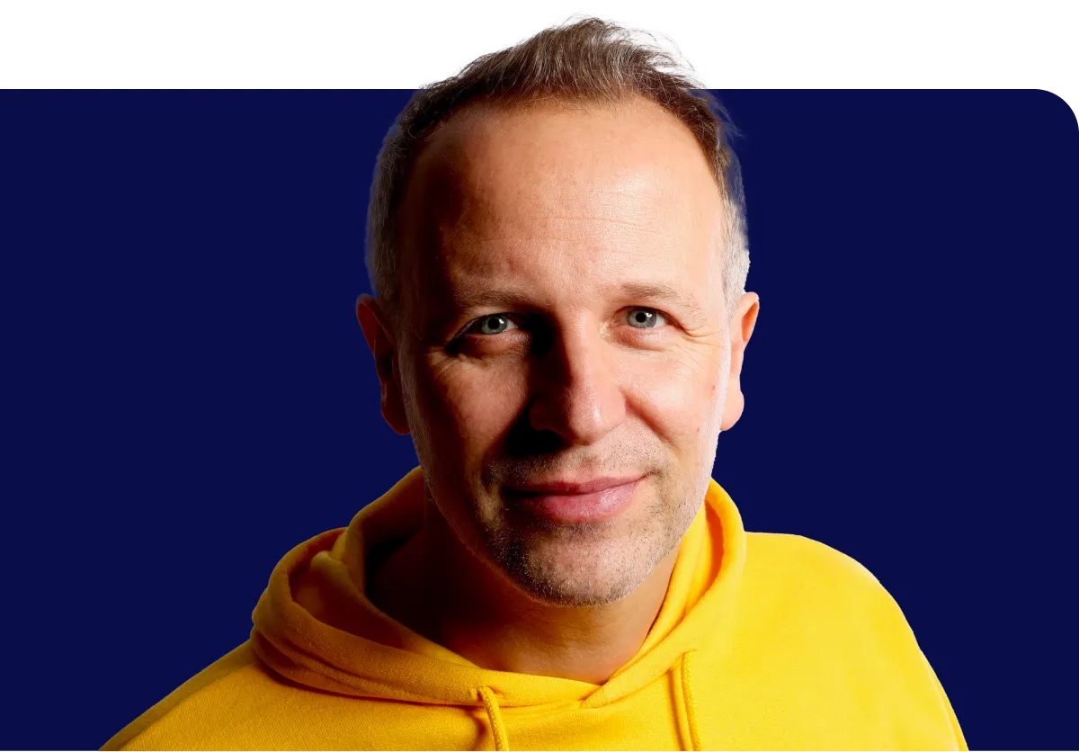 Man in a yellow hoodie smiling against a dark blue background.