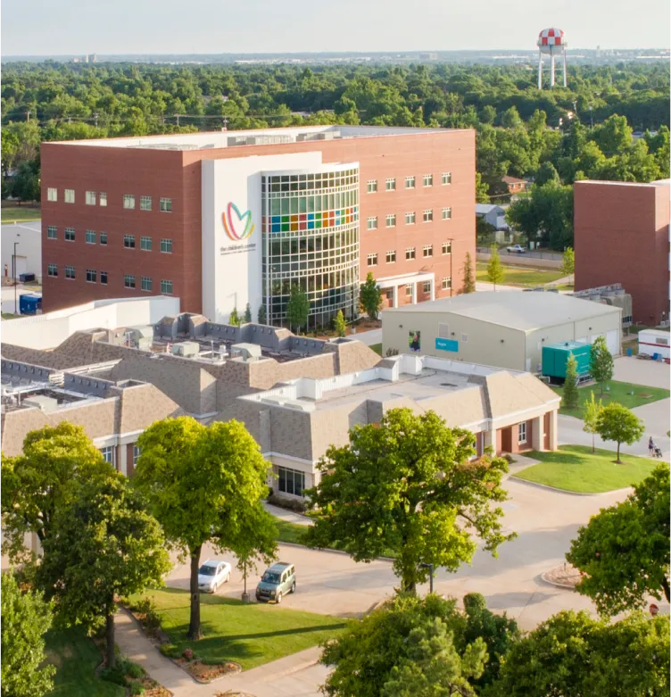 Bethany Children's Health Center's. Aerial view of a healthcare facility with red brick buildings, a large glass entrance, surrounding trees, and a distant water tower.