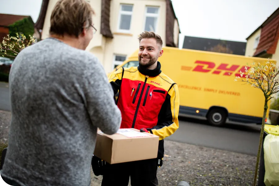 A DHL courier in a red and yellow uniform hands a package to a person outside a house, with a DHL van parked in the background.