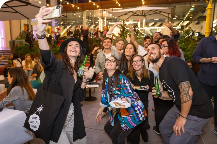 A group of people smiling and taking a selfie at a lively indoor gathering, with drinks and snacks, under string lights and umbrellas.