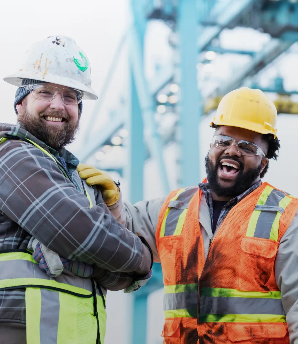 Two construction workers in safety gear smiling and laughing, with a blurred industrial background. One wears a white helmet, the other a yellow one.