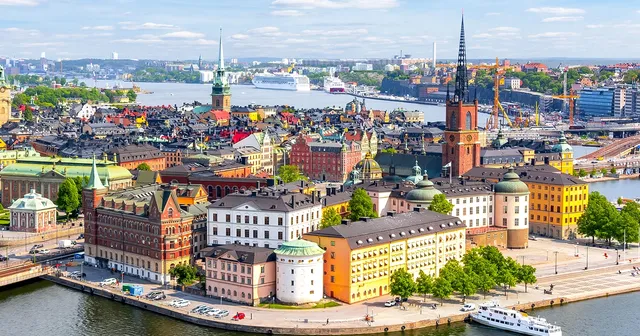 Aerial view of Stockholm's Gamla Stan with colorful historic buildings, water, boats, and a spire set against a partly cloudy sky.
