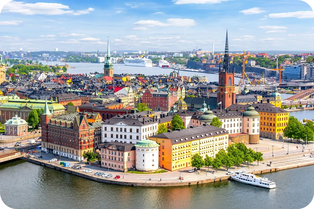 Aerial view of Stockholm's Gamla Stan with colorful historic buildings, water, boats, and a spire set against a partly cloudy sky.