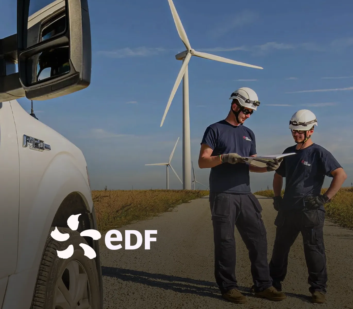 Two wind turbine technicians in hard hats review a clipboard beside a service truck, with large wind turbines in the background and the EDF logo overlaid
