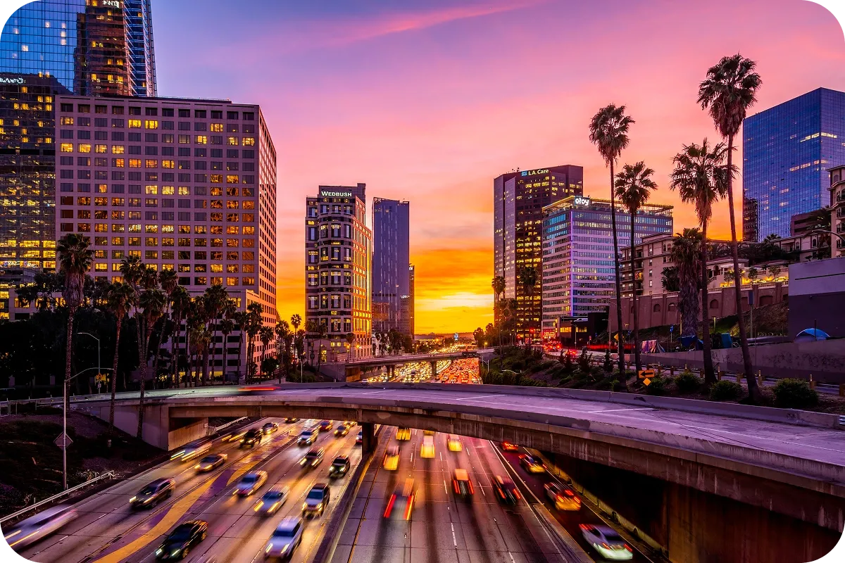 A vibrant cityscape at sunset with blurred traffic on a busy highway, surrounded by tall buildings and palm trees under a colorful sky.