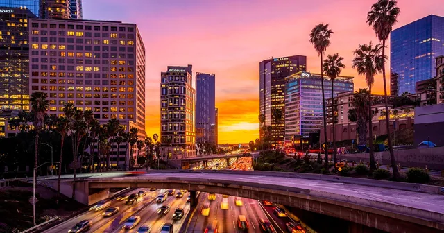 A vibrant cityscape at sunset with blurred traffic on a busy highway, surrounded by tall buildings and palm trees under a colorful sky.