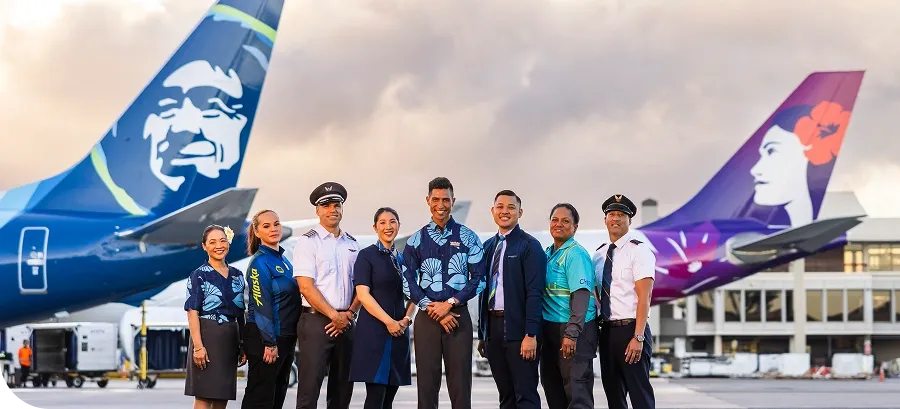 Airline crew members in uniforms stand in front of two airplanes with distinctive tail designs at an airport, under a cloudy sky.