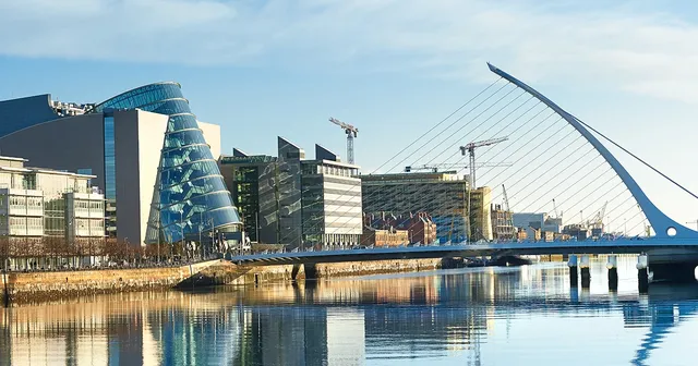 Modern cityscape with a river, glass buildings, cranes, and a distinctive white bridge reflecting in the calm water under a clear blue sky.