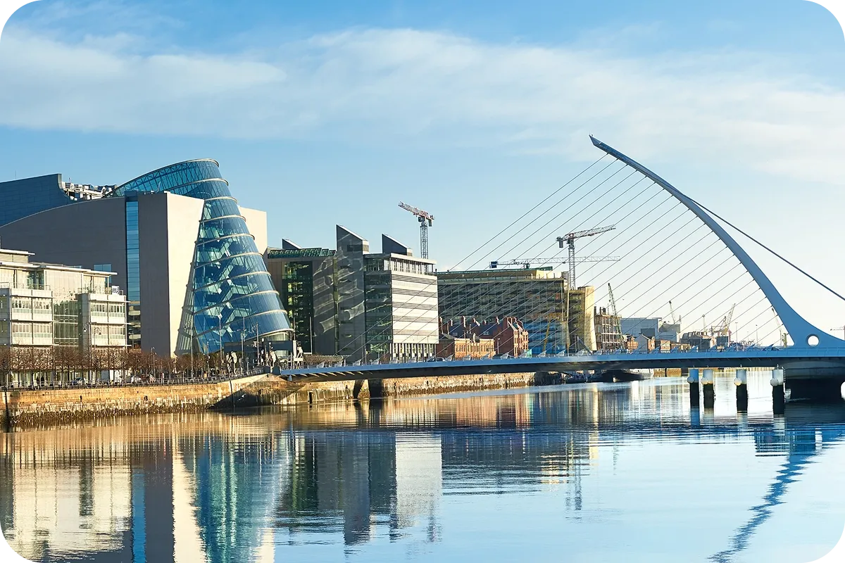 Modern cityscape with a river, glass buildings, cranes, and a distinctive white bridge reflecting in the calm water under a clear blue sky.