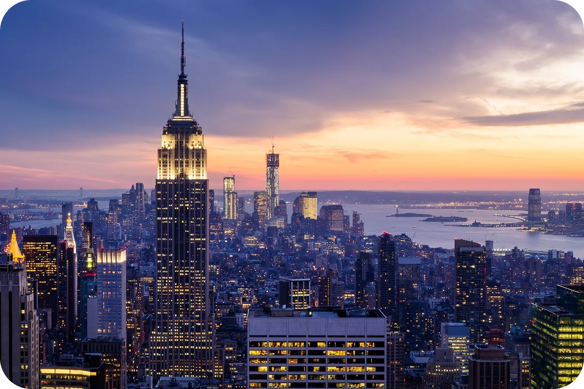 City skyline at dusk with illuminated skyscrapers, including the Empire State Building, against a colorful sunset sky and distant water view.