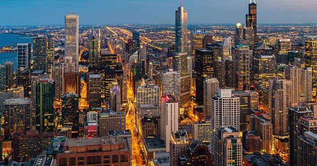 Aerial view of a cityscape at dusk, featuring illuminated skyscrapers, streets, and a vast expanse of buildings extending to the horizon.