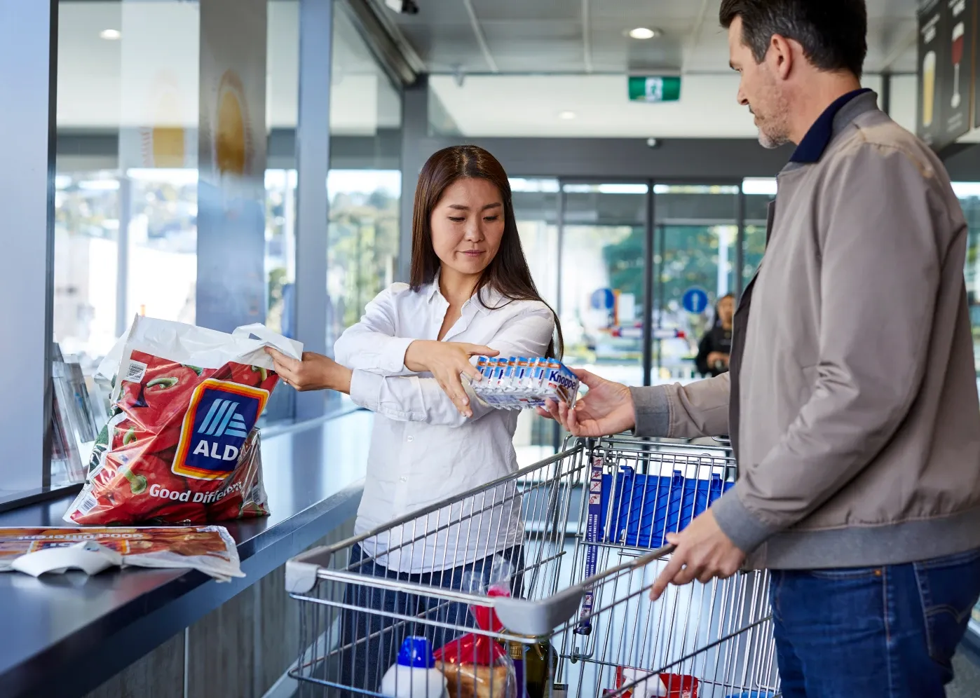 A woman and a man at a grocery store checkout, placing items into a shopping cart. The woman holds a water bottle, and an Aldi bag is visible.
