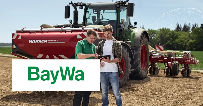 Two people in a field using a tablet in front of a red tractor. The BayWa logo is visible in the foreground.