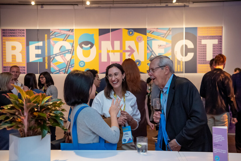 Internal communicators network at the welcome party at VOICES 2022 in New York City. A group of three people talk animatedly, smiling, and laughing with drinks in hand. Behind them are several other groups of communicators with a multi-colored sign that says