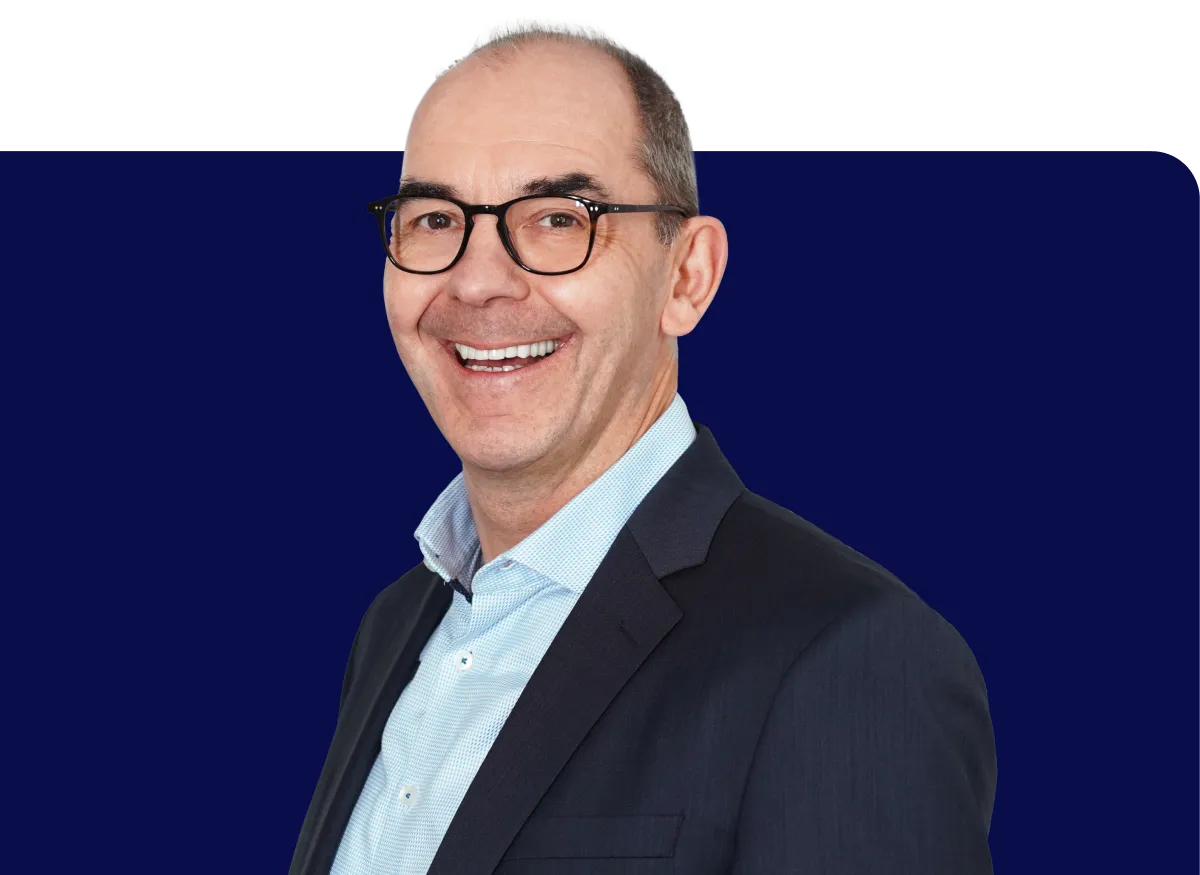 Smiling man in glasses and a dark suit with a light blue shirt, standing against a dark blue background.