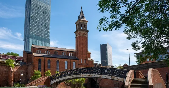 Historic brick church with a tall tower and arched bridge over a canal, set against modern skyscrapers under a clear blue sky.