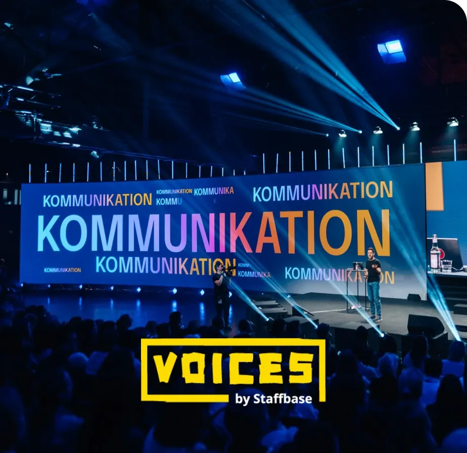 A speaker on stage at a conference with "Kommunikation" displayed in bold letters on a screen. The event is titled "Voices by Staffbase."
