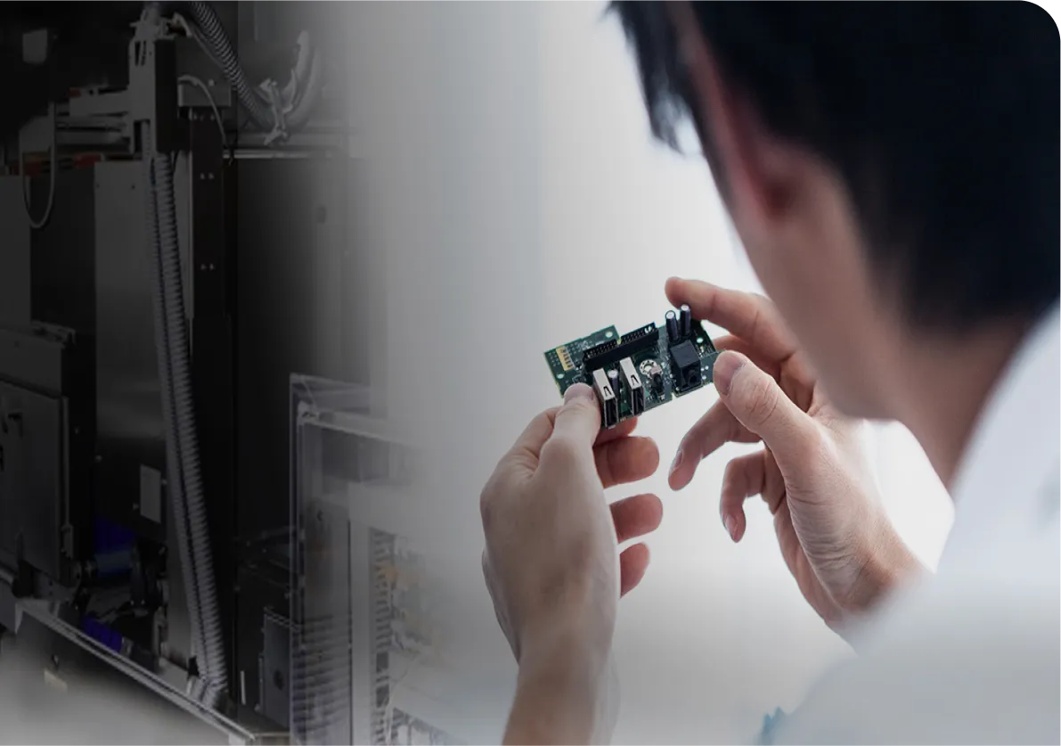 Person examining a small electronic circuit board, with blurred machinery in the background.