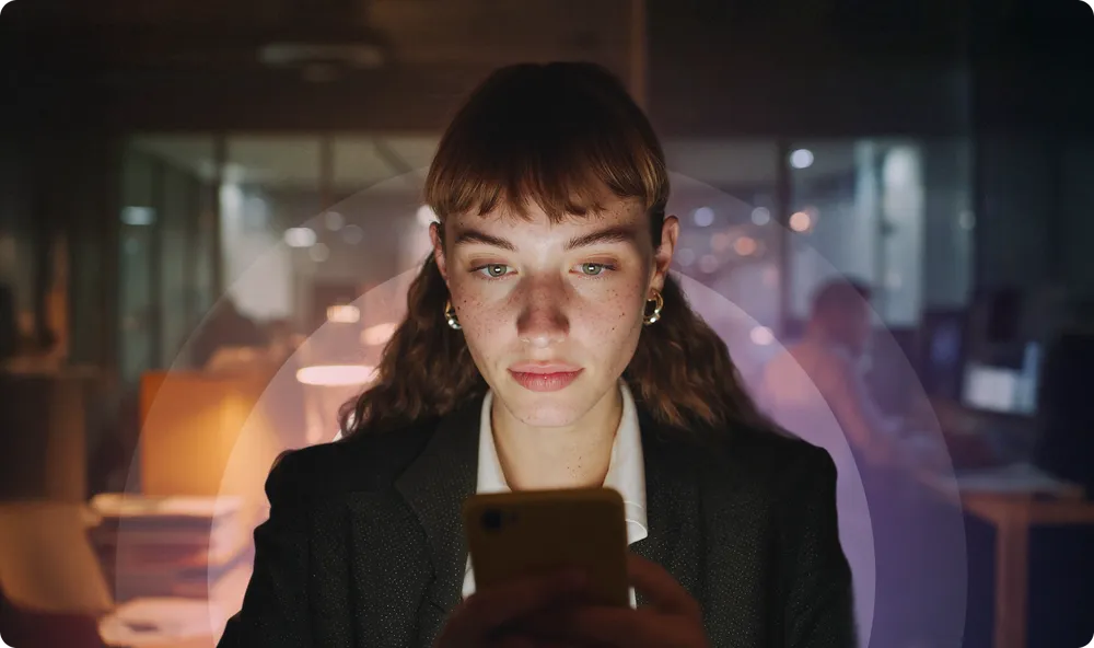 Woman in a dark office focused on her smartphone, with a soft light illuminating her face and blurred background.