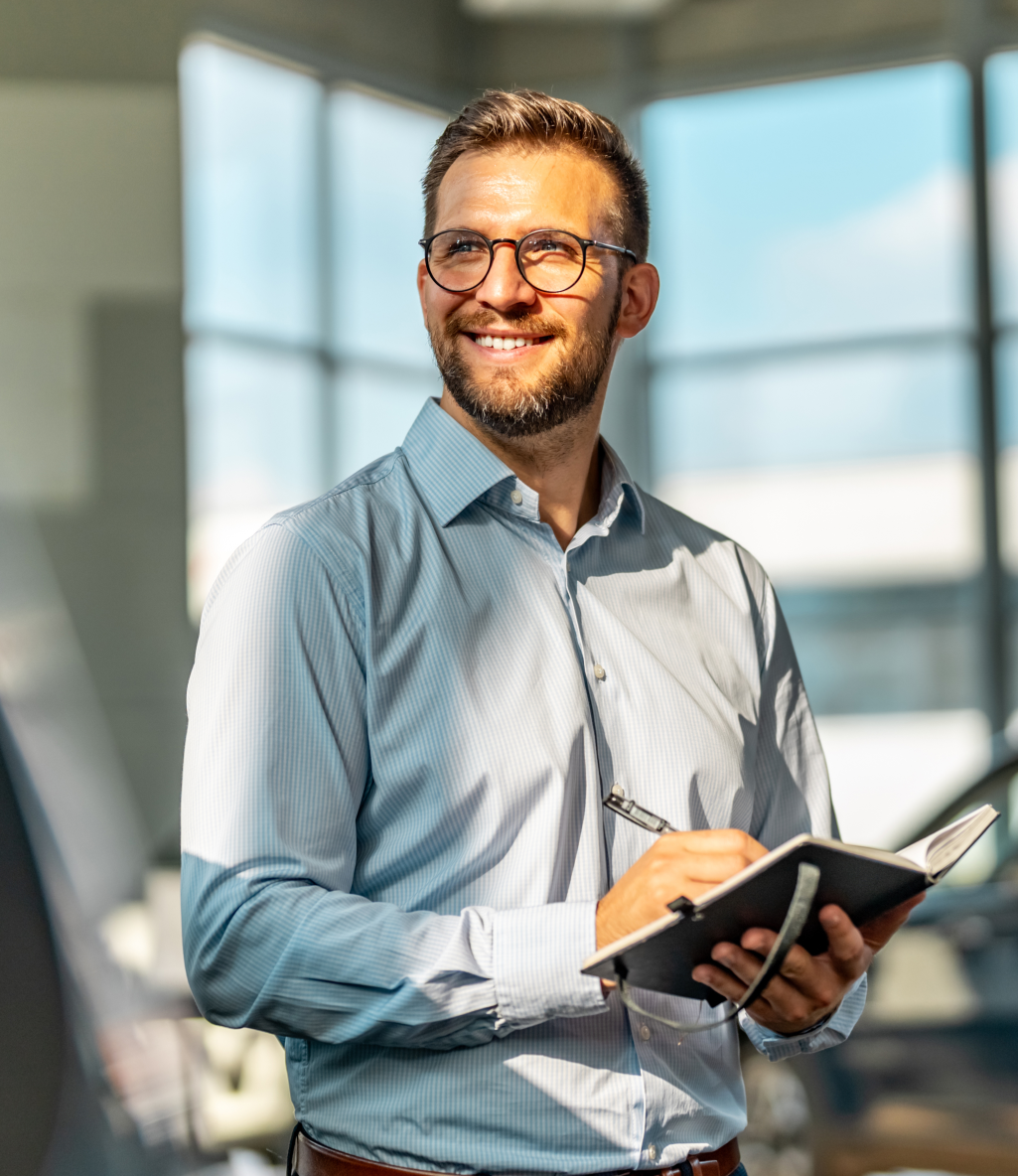 Smiling man with glasses and a beard, wearing a light blue shirt, holding a notebook and pen in a bright, modern office setting.