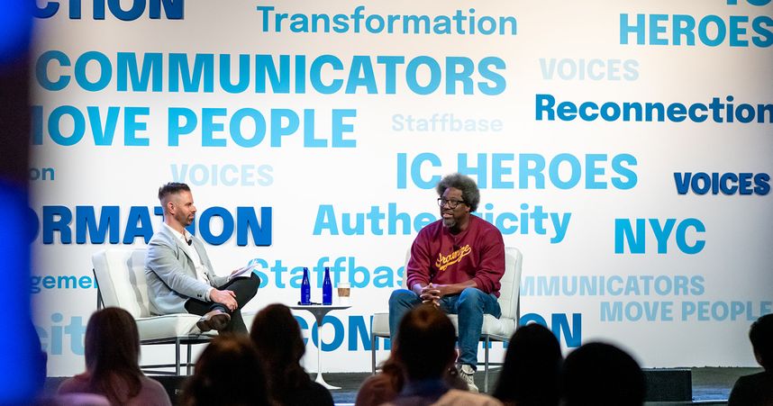 Comedian, author, and documentary producer W. Kamau Bell sits on stage with VOICES conference host Adam Brayford. They are on stage with words like "transformation" and "reconnection" and phrases like "communicators move people" in blue text behind them on a white background. We see the backs of the audiences' heads listening to Kamau and Adam.