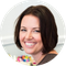 Smiling woman with short brown hair, wearing a dark top, sits in front of a blurred background with colorful objects.