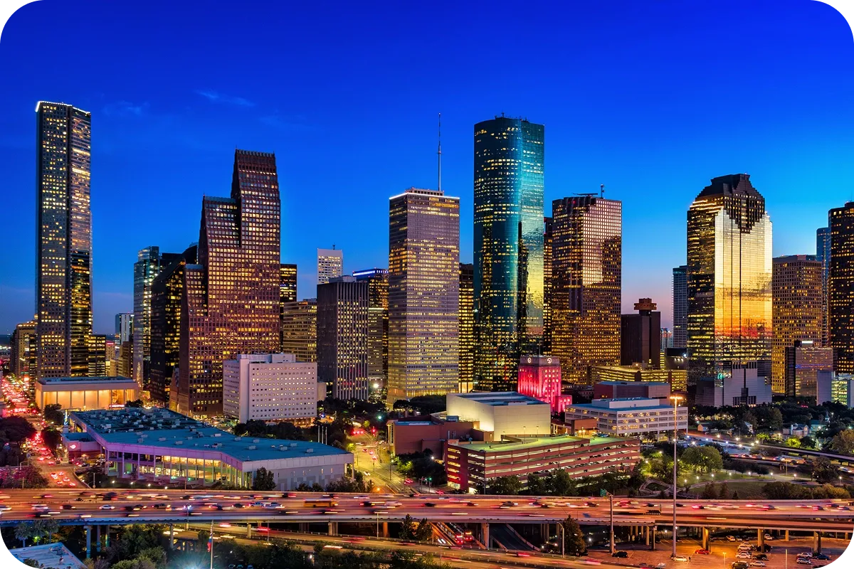 Skyline of a bustling city at dusk with illuminated skyscrapers, a vibrant blue sky, and busy highways in the foreground.