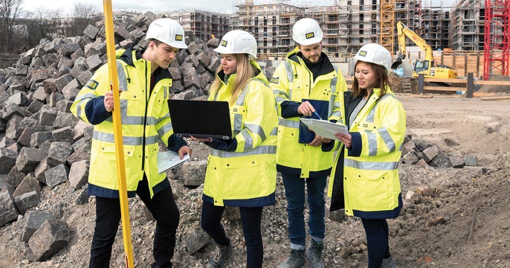 Four construction workers in yellow jackets and helmets discuss plans at a construction site, with rocks and buildings in the background.